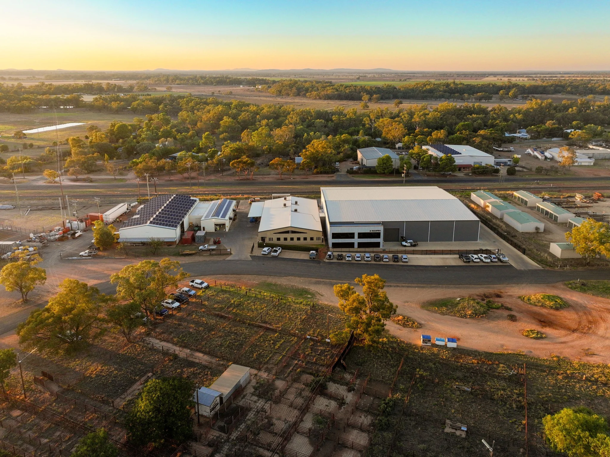 Aerial view of MASPRO facility complex with workshops, solar panels, parking, and surrounding farmland in daylight.