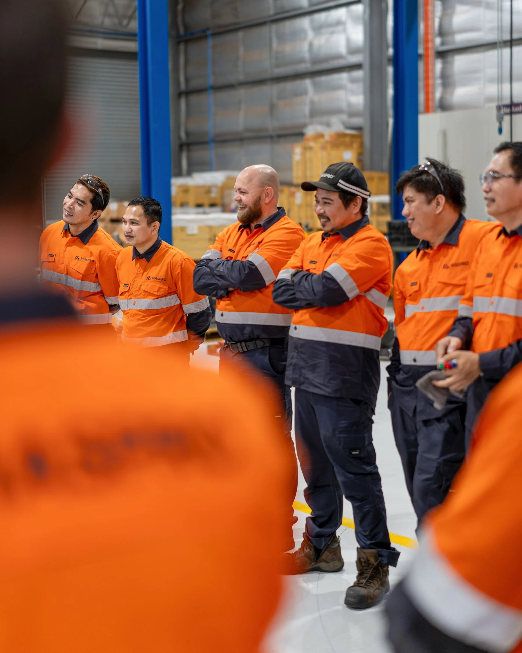 Group of six MASPRO technicians in orange hi-vis gathered together in a workshop, smiling and conversing.