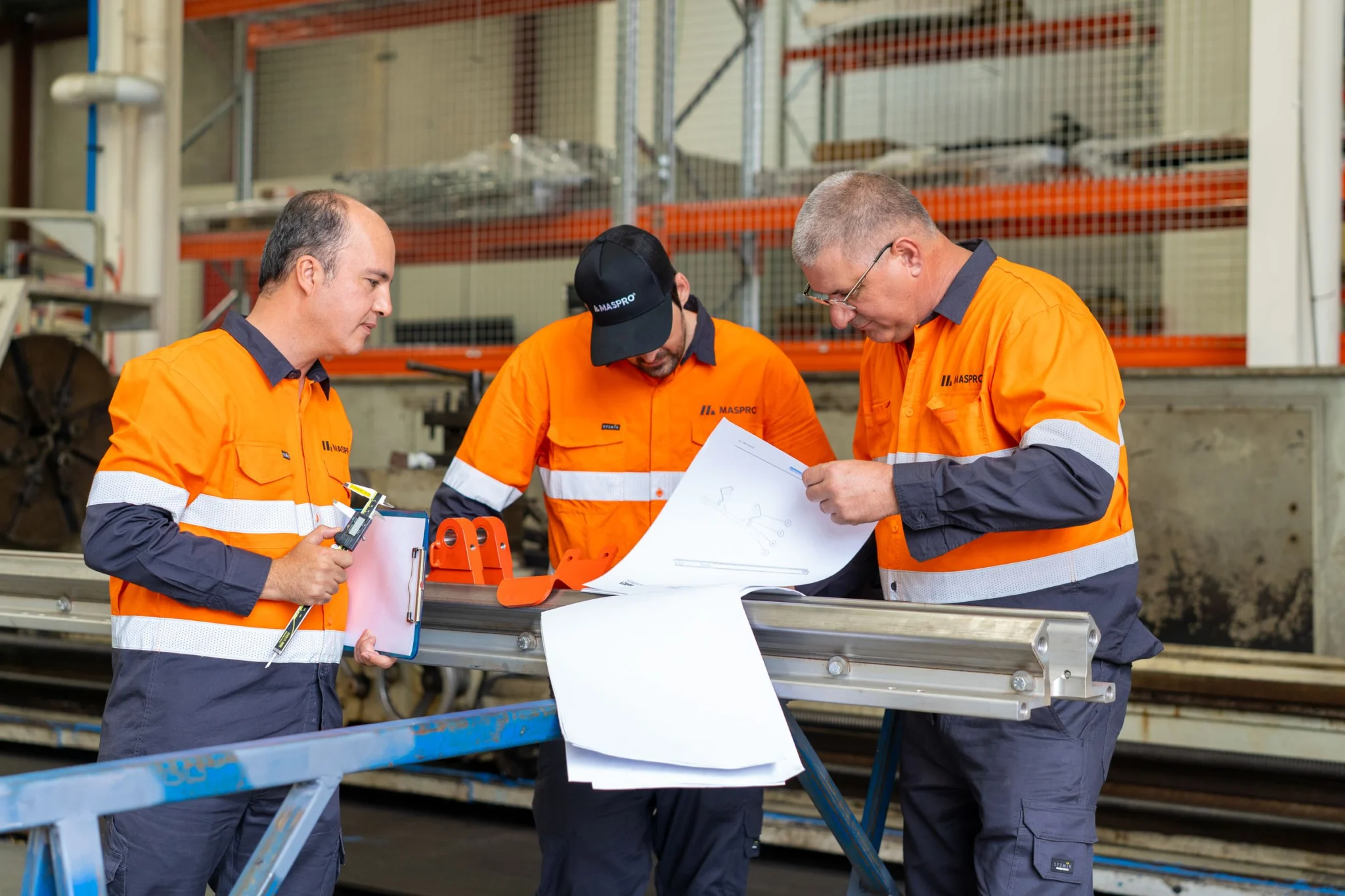 Three MASPRO technicians in orange hi-vis reviewing engineering blueprints and technical plans at a metal workbench.