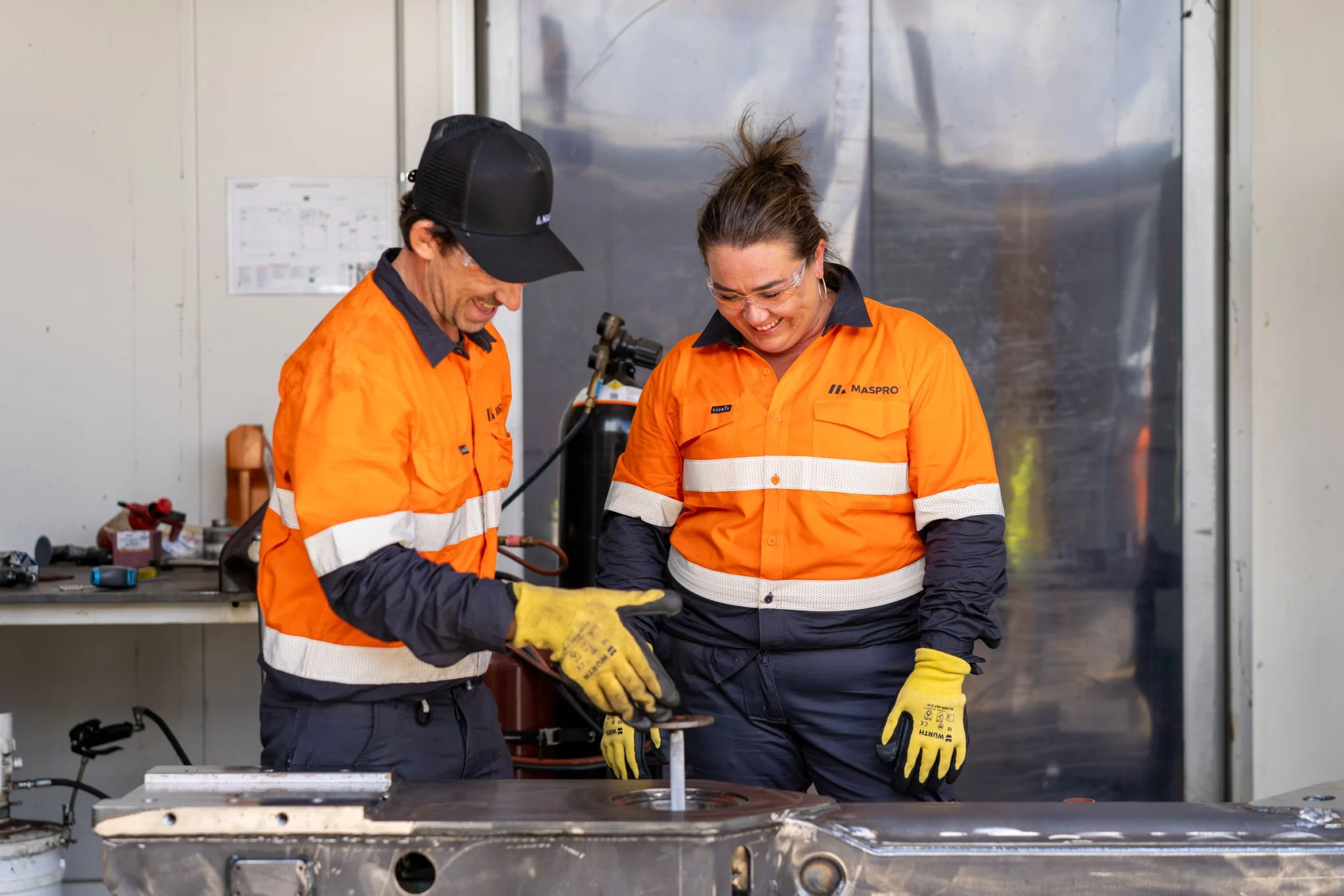 Two MASPRO technicians in yellow work gloves performing metal fabrication or assembly work on metal stock at a workbench.